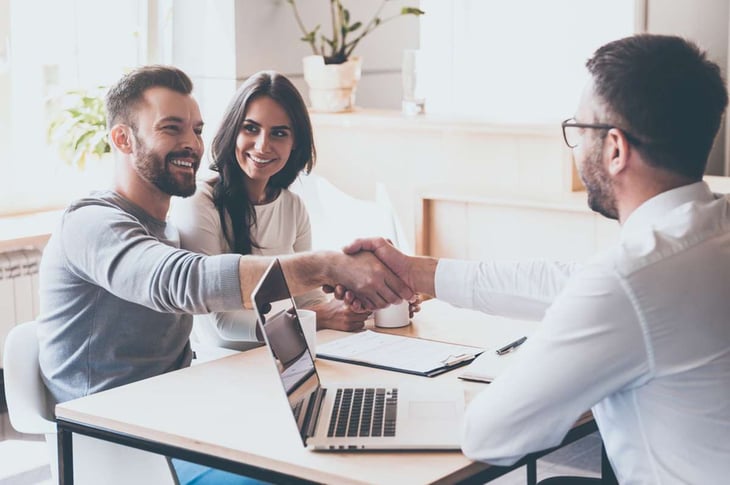 Cheerful young man bonding to his wife while shaking hand to man sitting in front of him at the desk