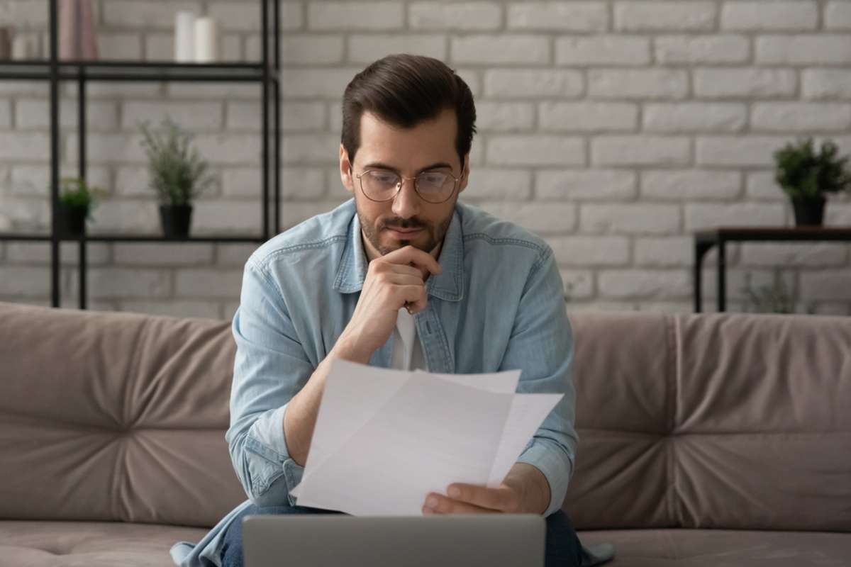 Pensive Caucasian young male worker sit on sofa at home work on laptop consider read post paper documents